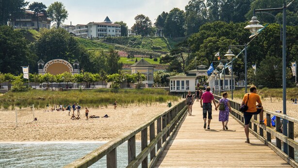 Um cais de madeira que leva a uma praia de areia, com pessoas caminhando e sentadas.