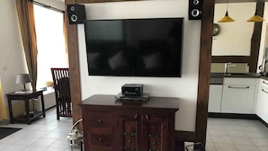 A living room with a flat-screen TV mounted on a wall, a wooden cabinet below, a small table with a lamp, and a kitchen area with cabinets and a sink. - Holiday flat at the market (Gingst)