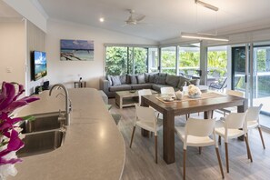 A modern kitchen with a central island, a dining area with a wooden table and white chairs, a grey sofa, and a large window with a view of greenery. - Oasis 12 & Free Buggy - Hamilton Island (Hamilton Island)