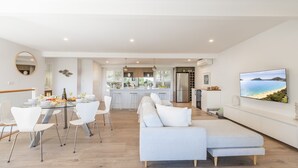Una cocina moderna con una isla central, un comedor con sillas blancas, un sofá y un televisor que muestra una escena de playa.
