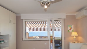 Ein Schlafzimmer mit Bett, Nachttisch, Lampe, Fenster mit Aussicht, Deckenventilator und Tür zu einem Außenbereich.