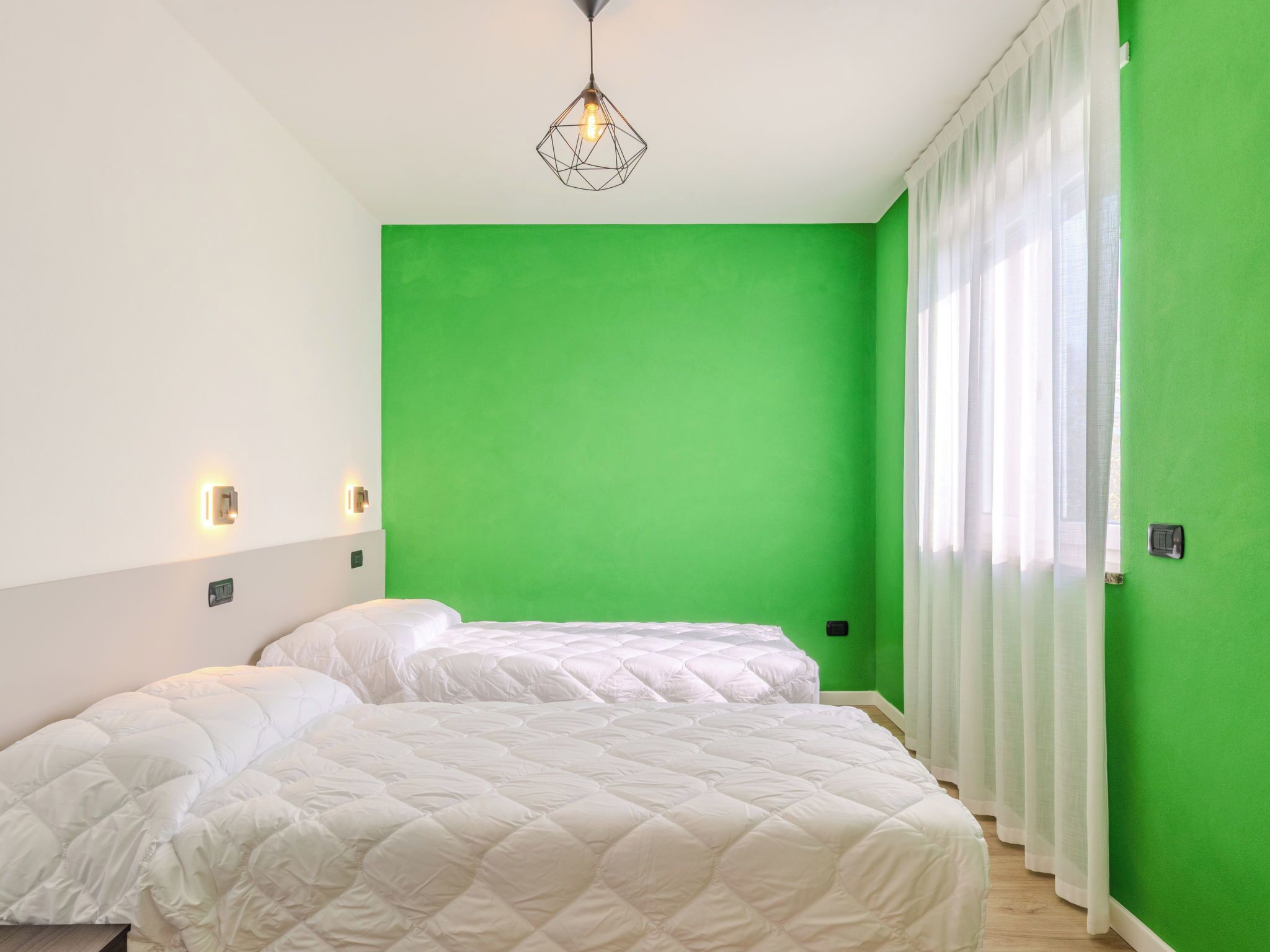 A room with two beds, a green wall, a hanging light fixture, and a window with sheer curtains.