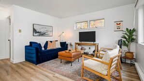 A living room with a blue sofa, a wooden coffee table, a TV, and a large window with artwork above.