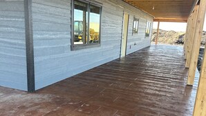 A covered wooden deck with a grey exterior wall and large windows.