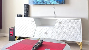 Uma sala de estar com uma TV de tela plana na parede, um console branco, uma mesa de centro redonda com um jogo americano vermelho e um controle remoto sobre a mesa.