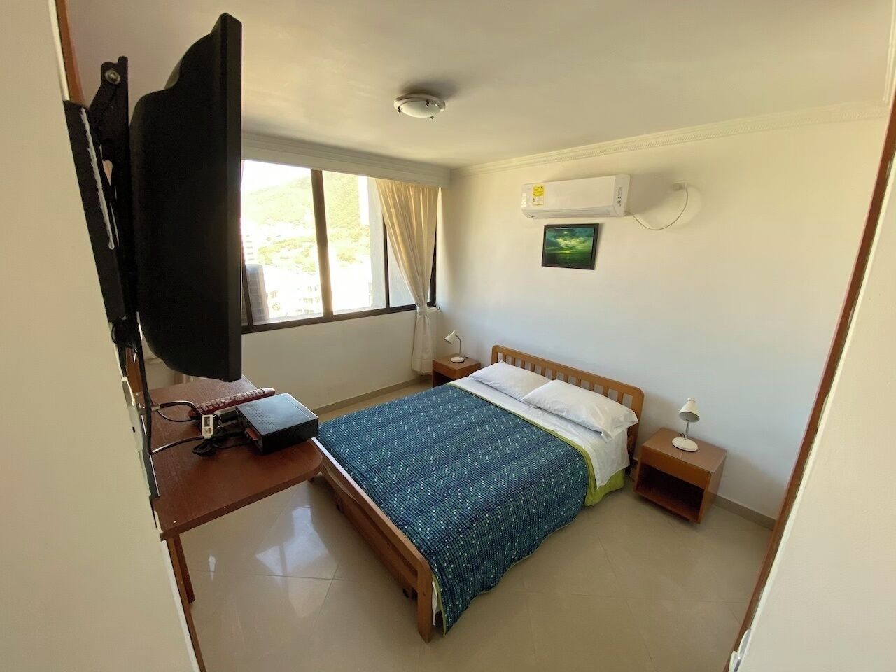 Quarto com cama, mesas de cabeceira, escrivaninha com computador, televisão e janela.