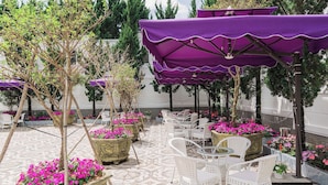 Un espace extérieur aménagé avec des chaises blanches et des parasols violets, entouré de plantes et de fleurs en pot.