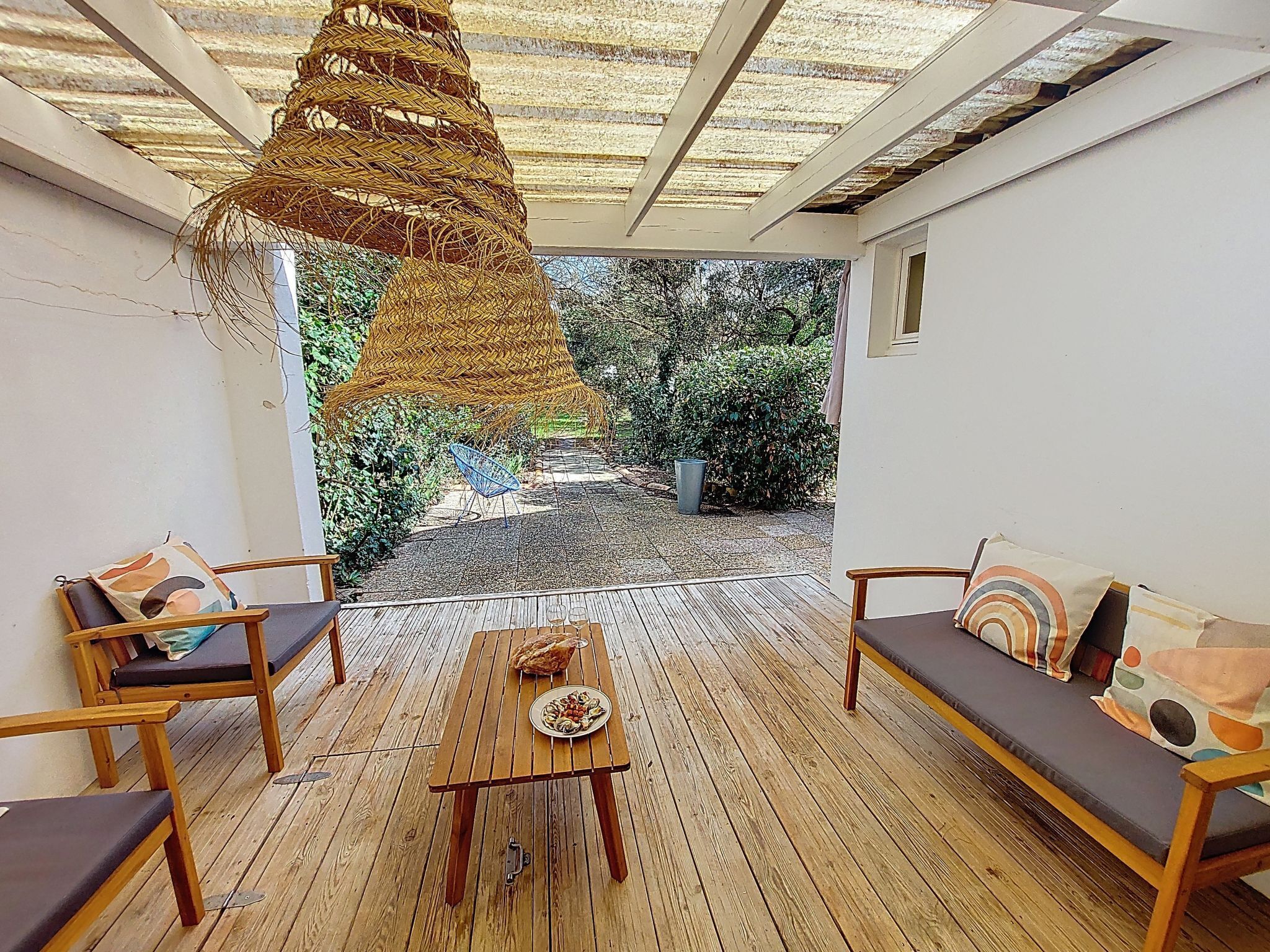 A covered patio with wooden flooring, two sofas, a table with a plate of food, and a hanging woven light fixture.