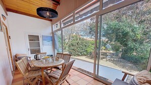 A dining area with a wooden table and chairs, a view of the garden through large windows, and a ceiling light fixture. - Villa Ecureuils by Interhome (Mimizan)