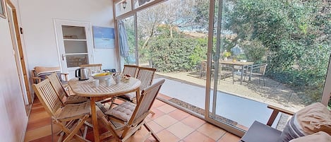 A dining area with a wooden table and chairs, a view of the garden through large windows, and a ceiling light fixture.