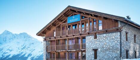 A multi-story wooden building with stone walls, labeled "LES BALCONS," featuring balconies and a mountainous backdrop.