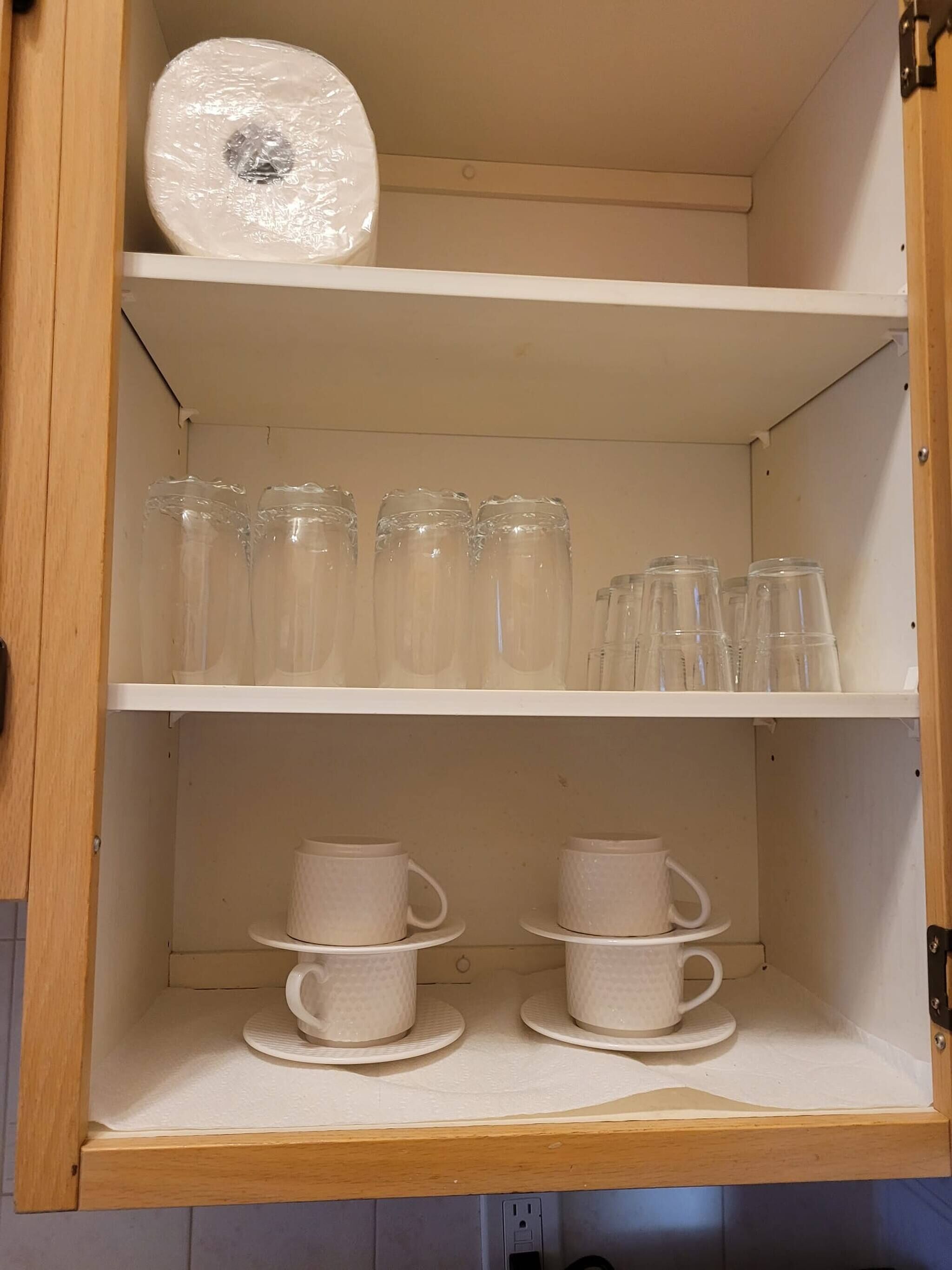 Un meuble en bois doté d’étagères où sont rangées des tasses en verre transparent, des tasses en céramique blanche et un rouleau d’essuie-tout.