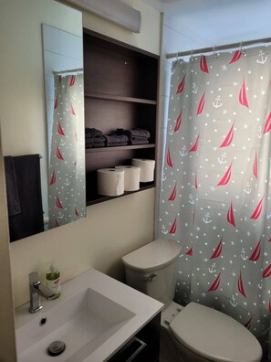 Une salle de bain avec un lavabo blanc, des toilettes, un miroir et une étagère contenant des serviettes et du papier toilette.