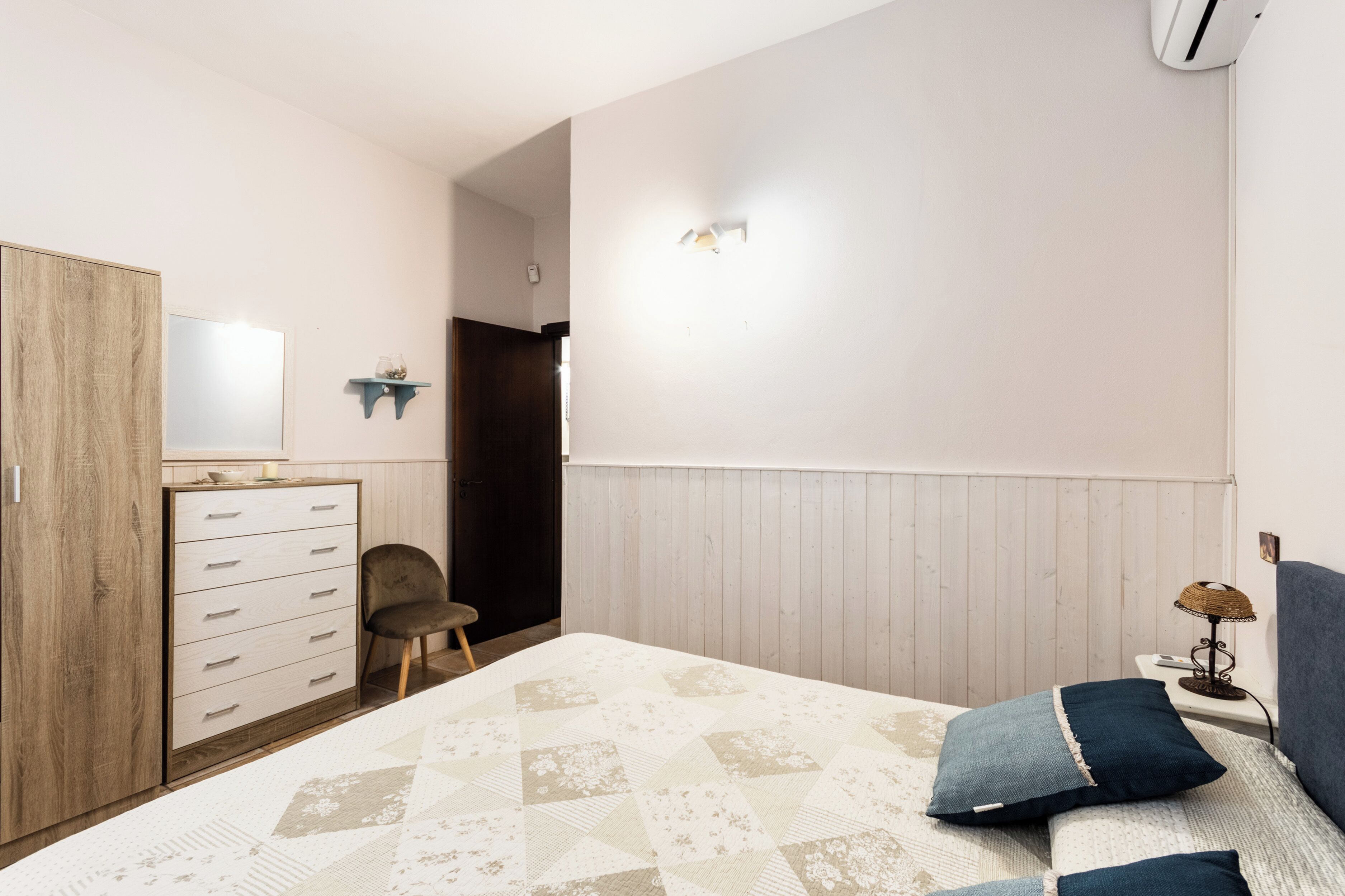 A bedroom with a bed, a dresser, a chair, a bedside table with a lamp, and a wall-mounted air conditioner.