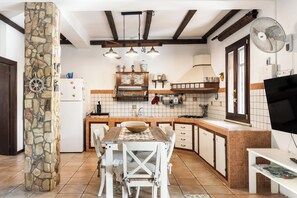 Une cuisine avec un pilier en pierre, des poutres en bois au plafond, une table à manger avec des chaises, une cuisinière encastrée et un téléviseur posé sur un meuble.