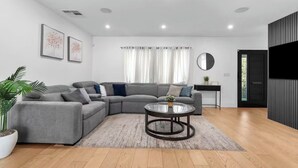 A modern living room with a grey sectional sofa, a glass coffee table, a potted plant, and a black TV unit. - 3BR Retreat w/ Pool & Spa, Prime Tarzana Location (Los Angeles)