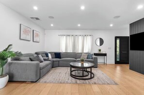 A modern living room with a grey sectional sofa, a glass coffee table, a potted plant, and a black TV unit. - 3BR Retreat w/ Pool & Spa, Prime Tarzana Location (Los Angeles)