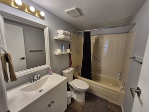 Een badkamer met een wastafel, toilet en bad. Er zijn planken met toiletartikelen en een douchegordijn.