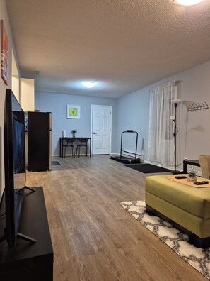 Uma sala de estar com televisão, sofá, mesa de jantar com cadeiras, uma porta e uma janela com cortinas.