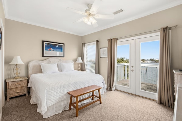 A bedroom with a bed, a nightstand, a bench, a ceiling fan, and a balcony with a view of the water.
