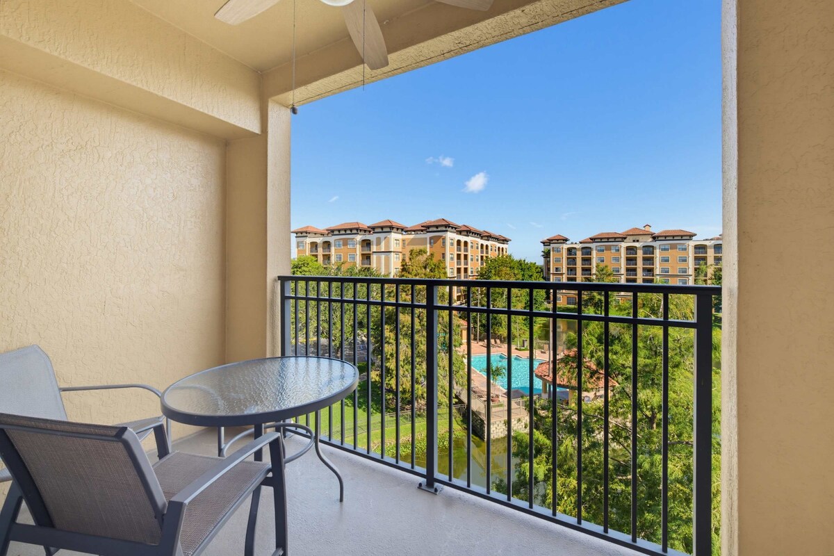 A balcony with a table and chairs, overlooking a swimming pool and apartment buildings.