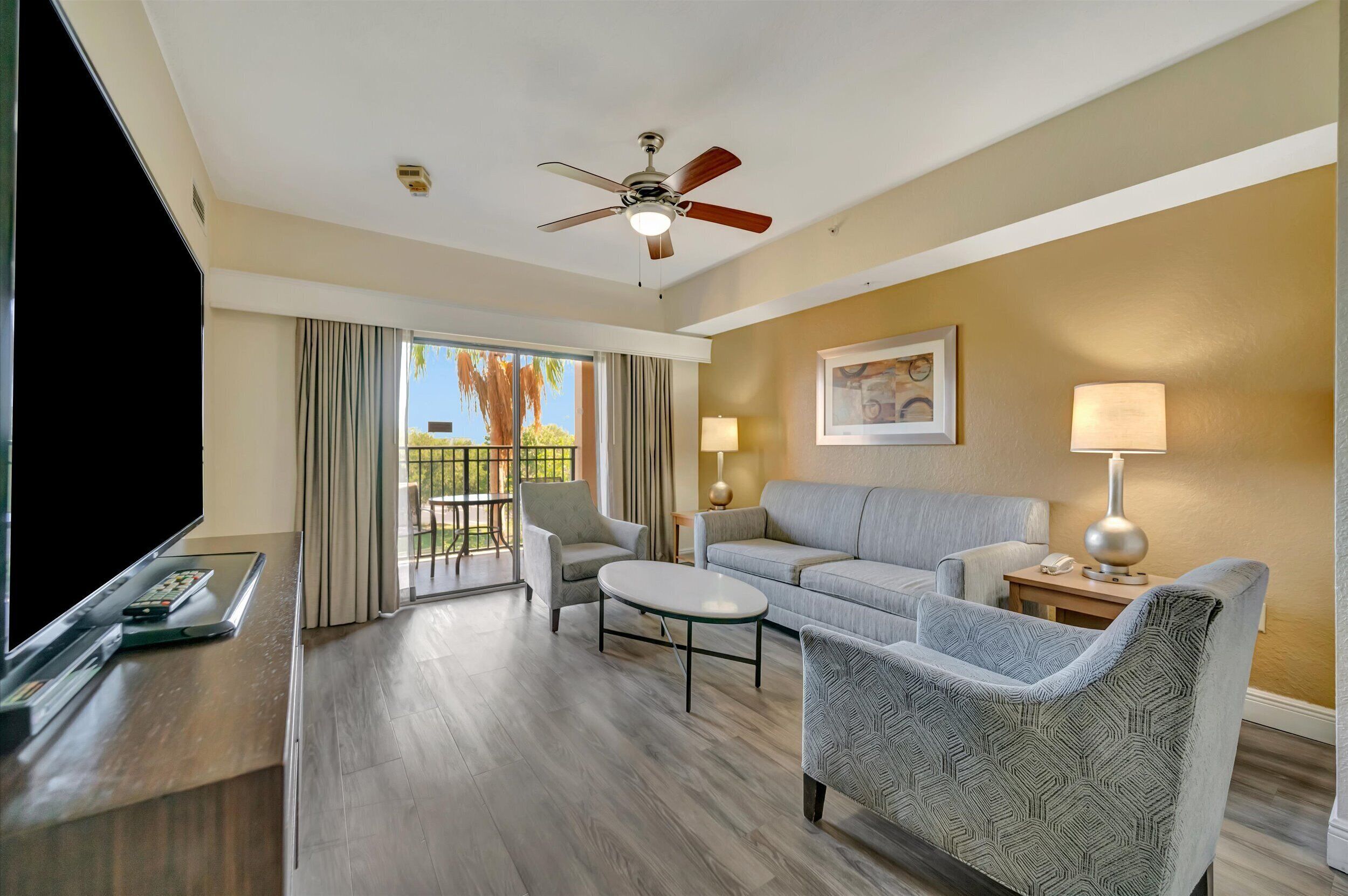 A hotel room with a flat-screen TV, a ceiling fan, a sofa, two armchairs, a coffee table, and a balcony with curtains.