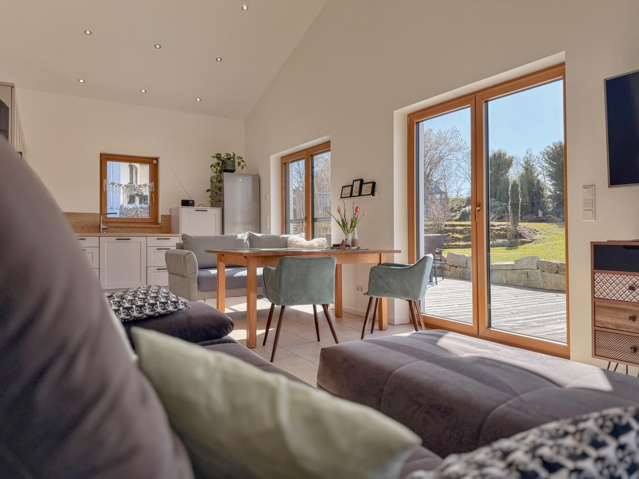 A modern living room with a sofa, dining table, and chairs. There is a kitchen area with a refrigerator and cabinets. Large windows provide a view of the outdoors.