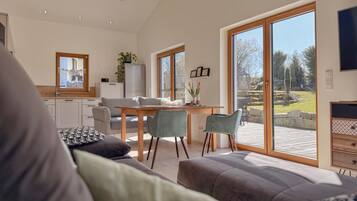 A modern living room with a sofa, dining table, and chairs. There is a kitchen area with a refrigerator and cabinets. Large windows provide a view of the outdoors.