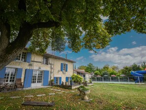 A rustic building with blue shutters, a large tree in the foreground, and a clear blue sky. - Spacious familyhouse with privat pool and slide (Lusignac)
