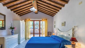 A bedroom with a wooden ceiling, a bed with a blue bedspread, a nightstand with a lamp, and a dresser with a vase of flowers. - Preselle by Interhome (Casale Marittimo)