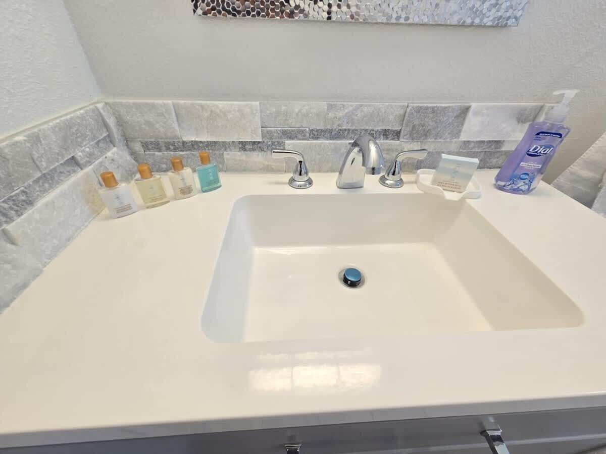 Un baño con un lavamanos blanco, dos grifos y un espejo. Hay artículos de aseo en la encimera.