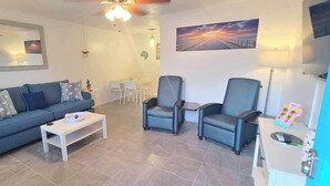 Ein Wohnzimmer mit einer Couch, zwei Sesseln, einem Couchtisch, einem Esstisch mit Stühlen, einem Deckenventilator und Kunstwerken an den Wänden.