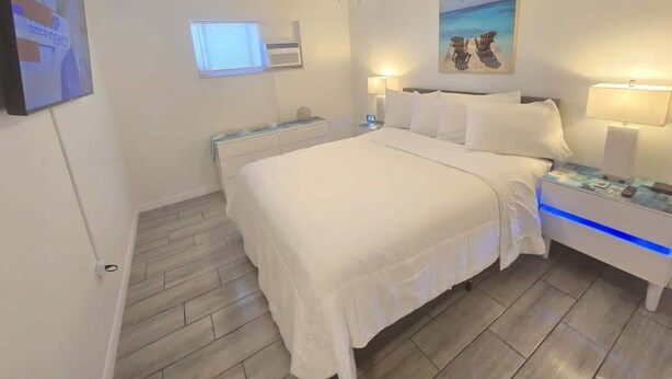 Ein Schlafzimmer mit Bett, Nachttischen, Deckenventilator, Fenster und Wandbild.