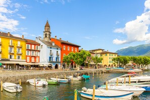 A waterfront with colorful buildings, a church tower, and boats docked along the shore. - Casa Viola Garden in Losone for 4 (Losone)