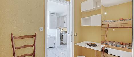 A compact room with a wooden chair, a small stool, a bunk bed, and a kitchenette in the background.