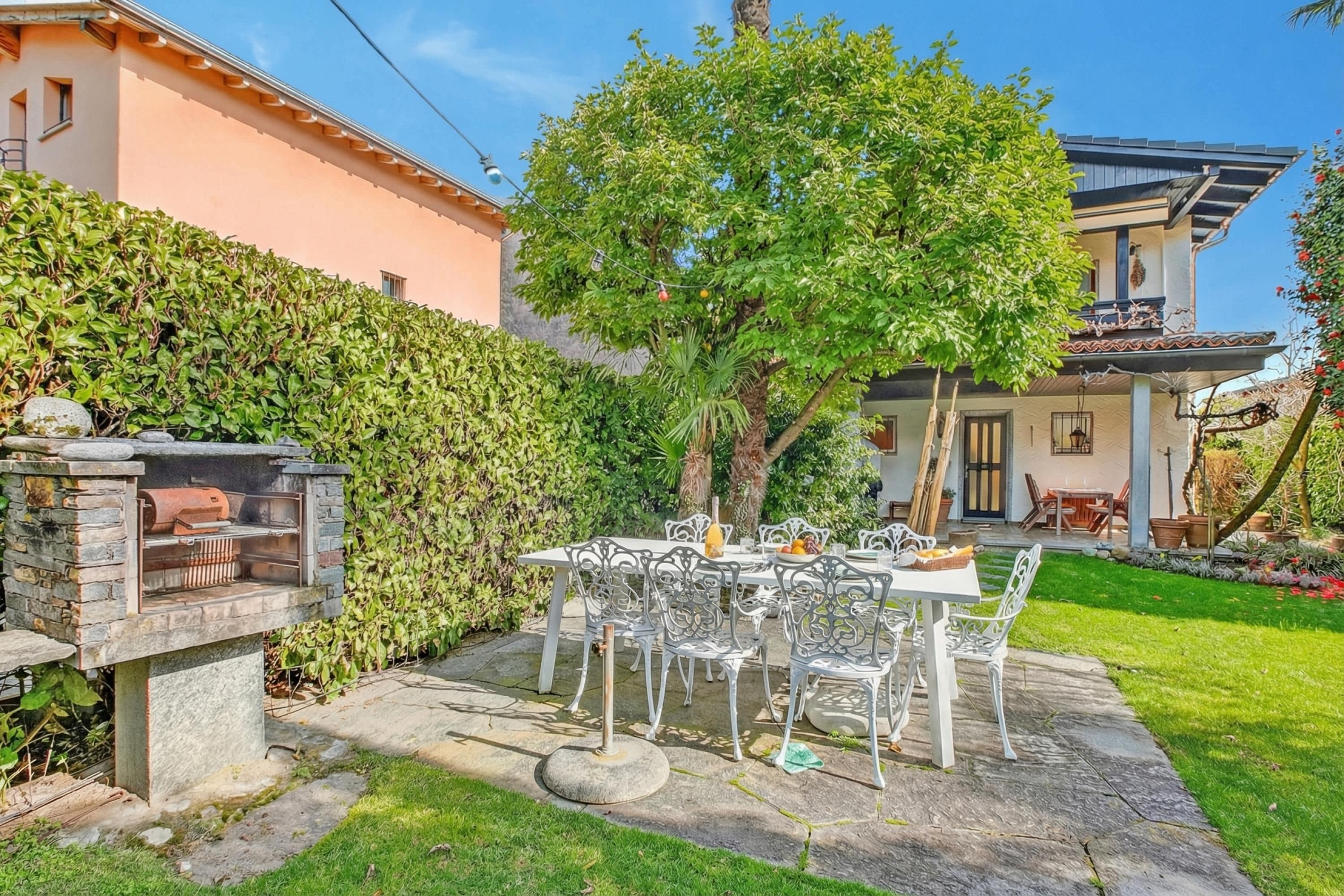 A patio with a stone barbecue grill, white metal chairs, and a table set for dining.