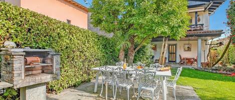 A patio with a stone barbecue grill, white metal chairs, and a table set for dining.