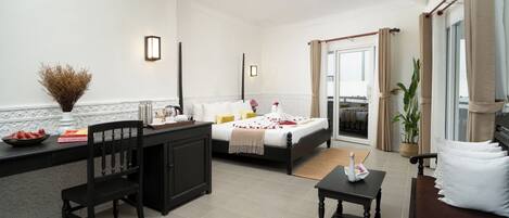 A hotel room with a bed, a desk, a chair, a small table, and a potted plant.