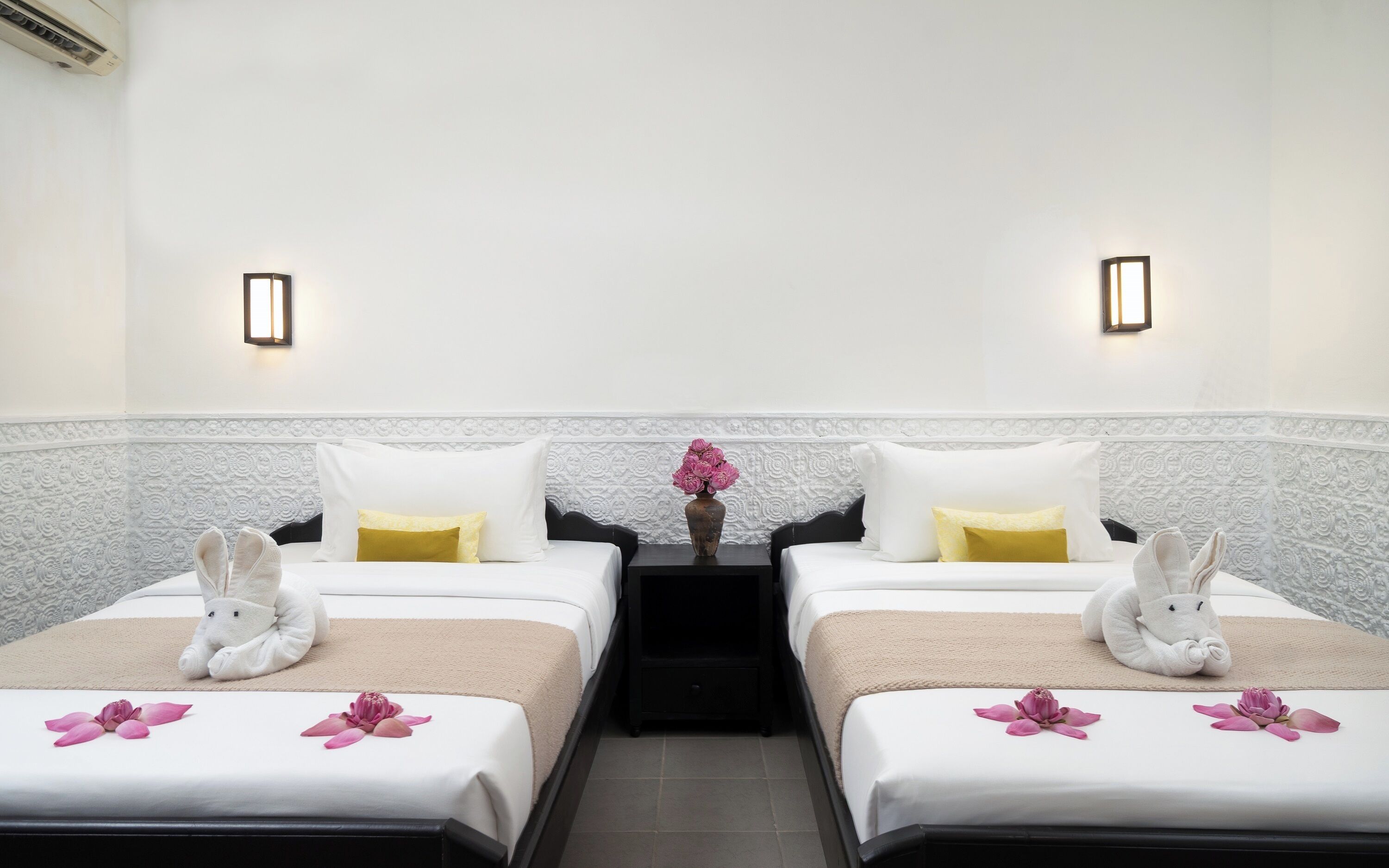 A hotel room with two beds, a nightstand, a vase with flowers, and a wall-mounted light fixture.
