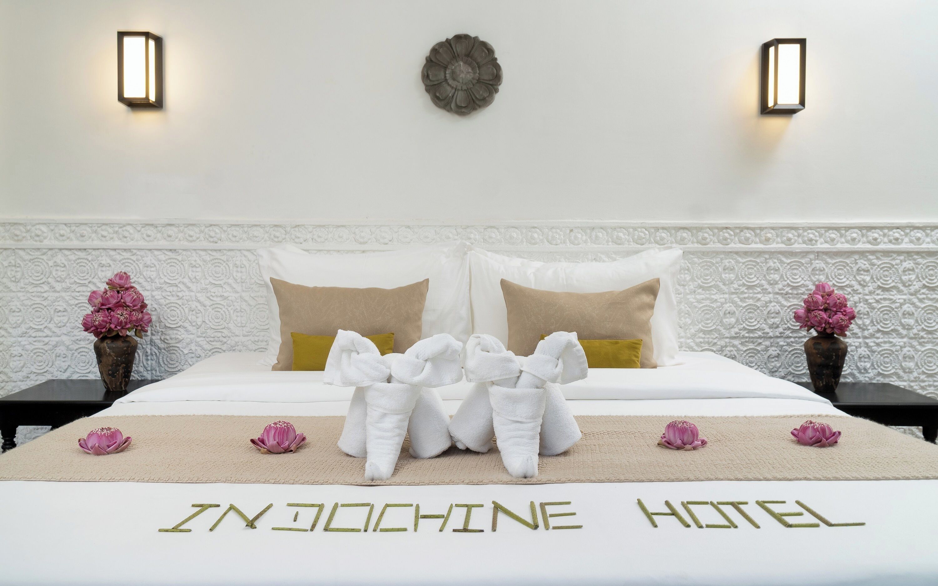 A hotel room with a bed, two nightstands, decorative pillows, a wall-mounted light fixture, and a floral arrangement on the nightstands.