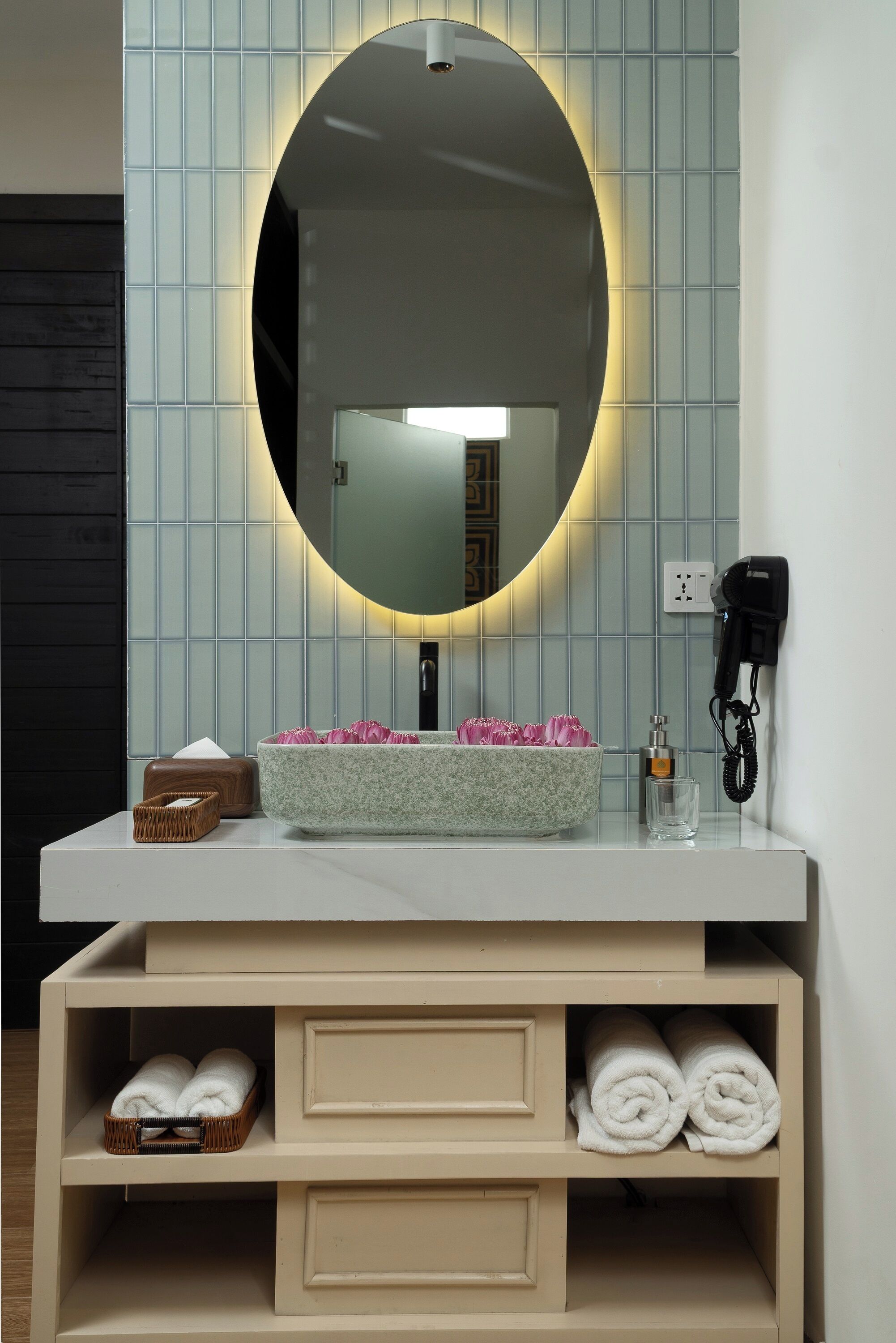 Une salle de bain avec un miroir rond, un lavabo orné d’un motif floral, un sèche-cheveux et des étagères pour ranger les serviettes.