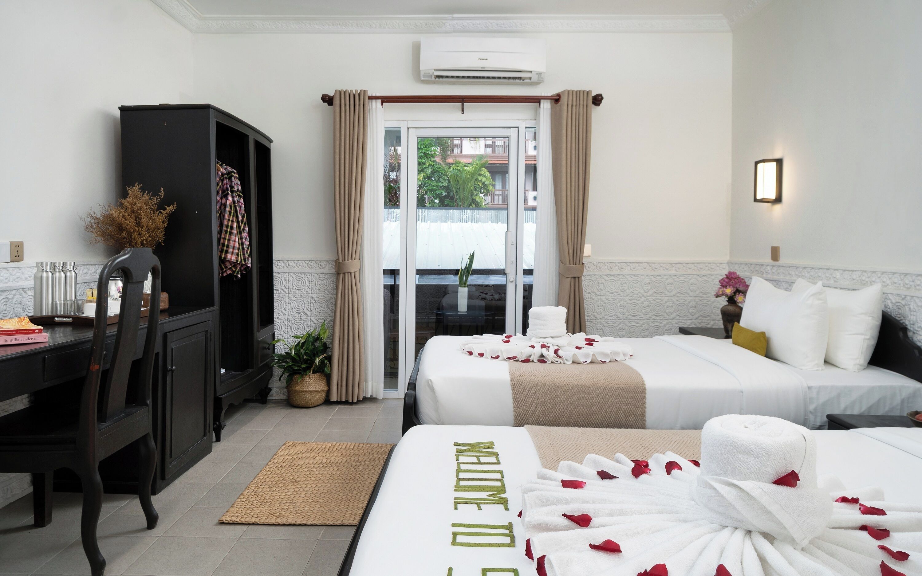 A hotel room with a bed, a desk, a wardrobe, a potted plant, and a view of the outside.