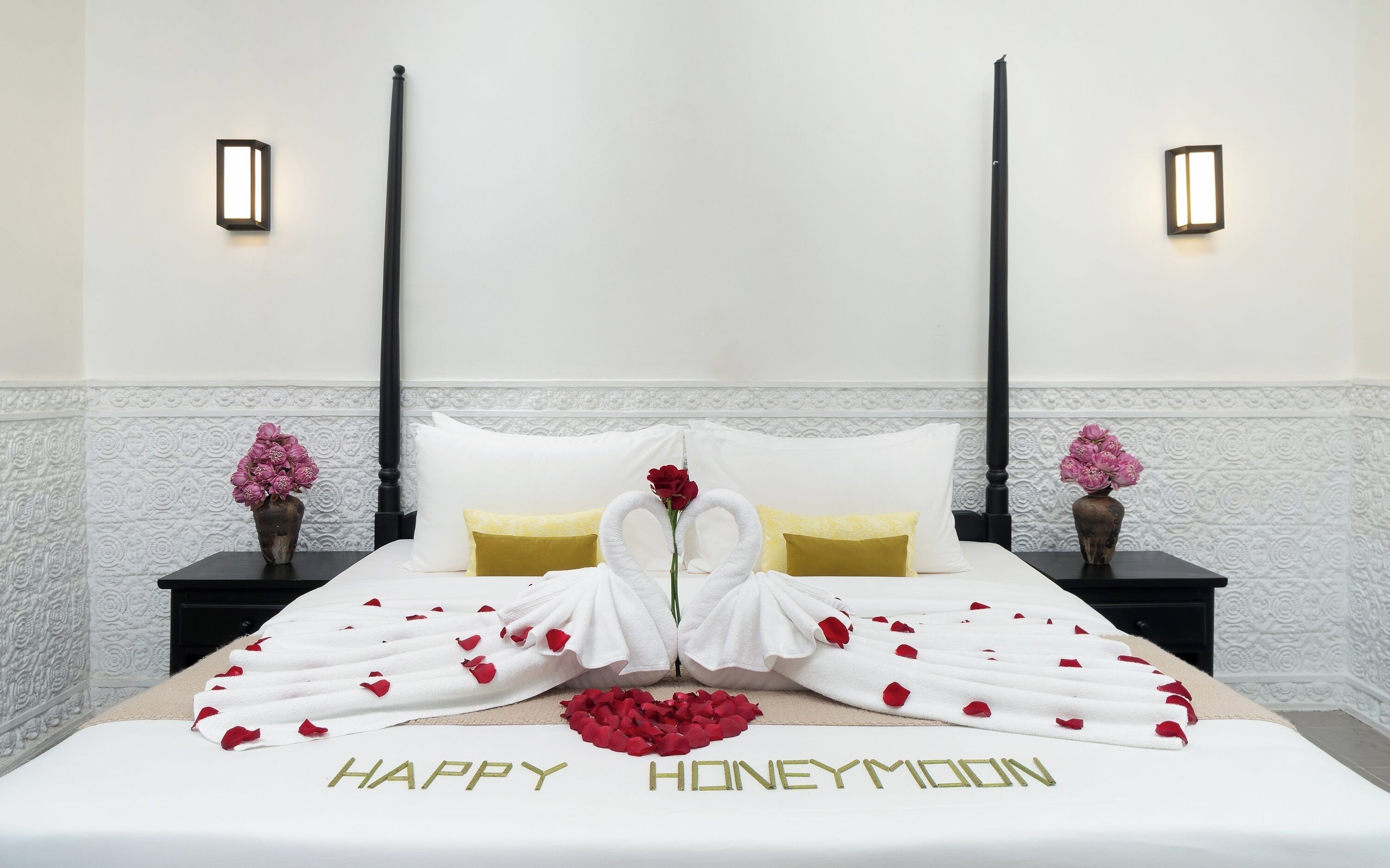 Una suite per la luna di miele con un letto decorato da asciugamani bianchi a forma di cigno, petali di rosa rossa e la scritta 'HAPPY HONEYMOON' sul copriletto.