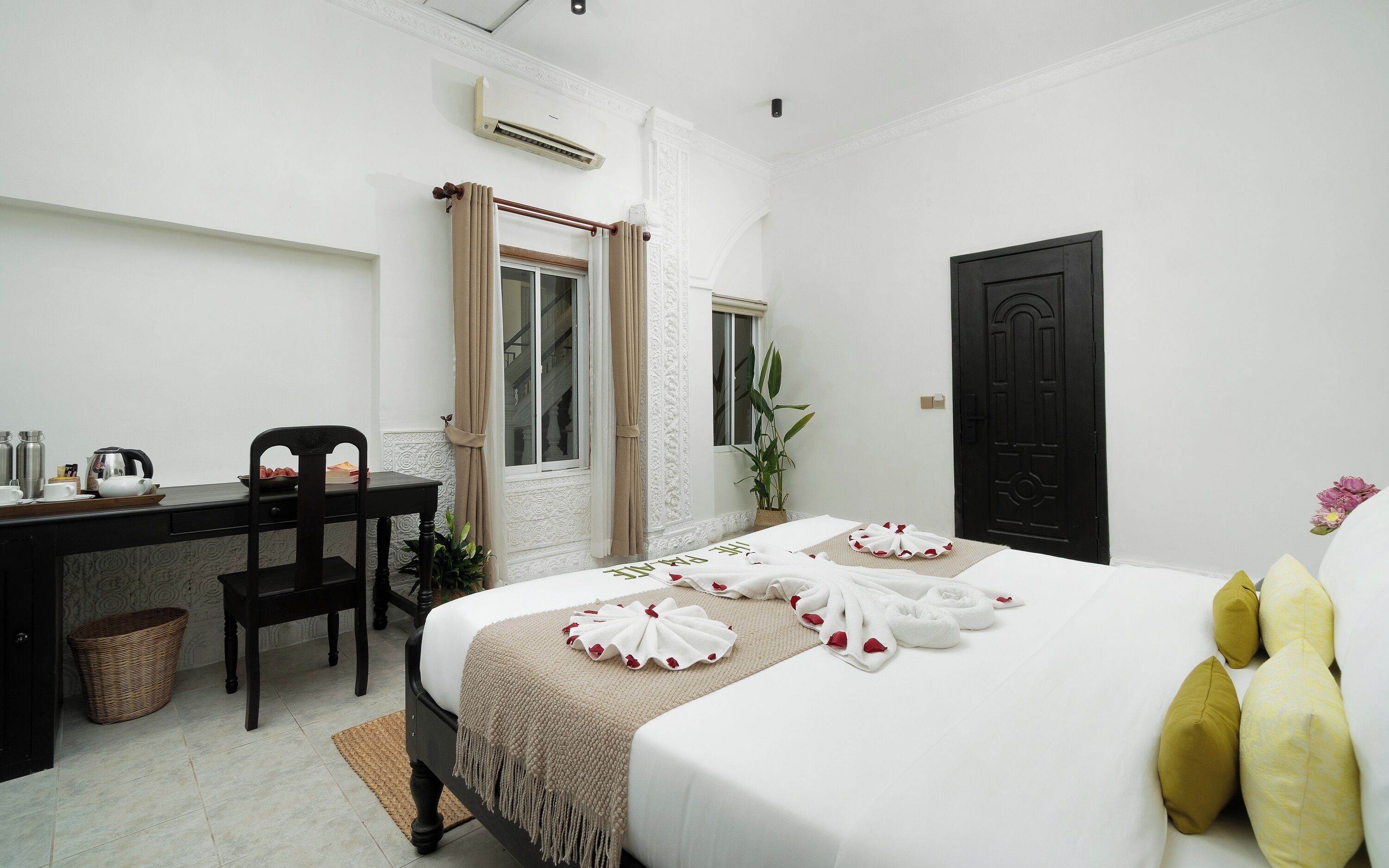 A hotel room with a bed, a desk, a chair, a window with curtains, a potted plant, and a black door.