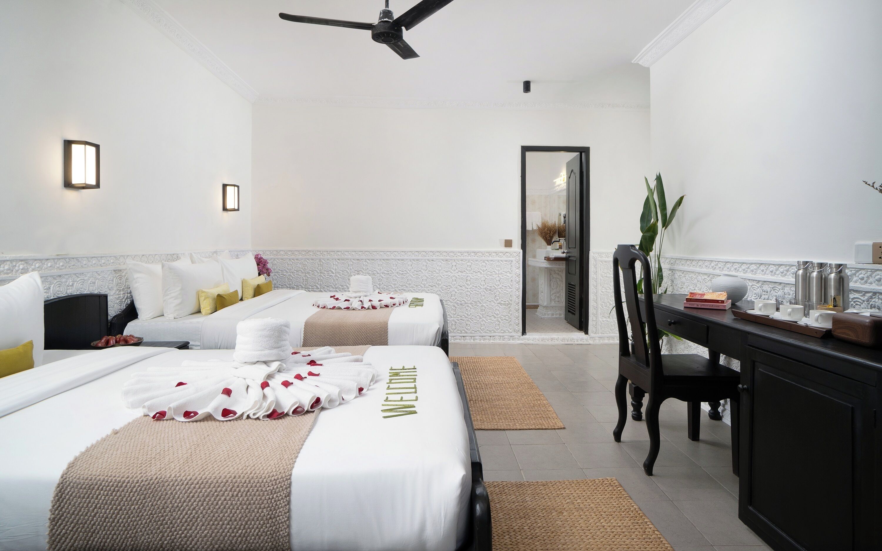 A hotel room with two beds, a desk, a chair, a ceiling fan, and a bathroom visible through an open door.