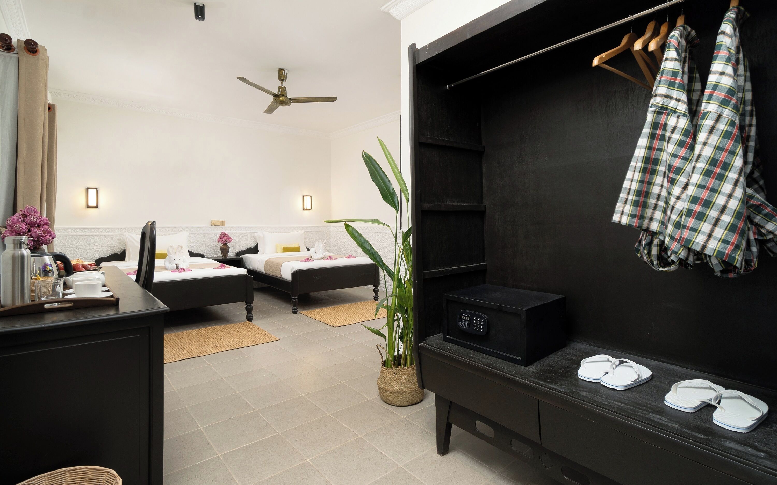 A hotel room with two beds, a desk with a tray of breakfast, a plant, a black cabinet, and a clothing rack with a plaid shirt.