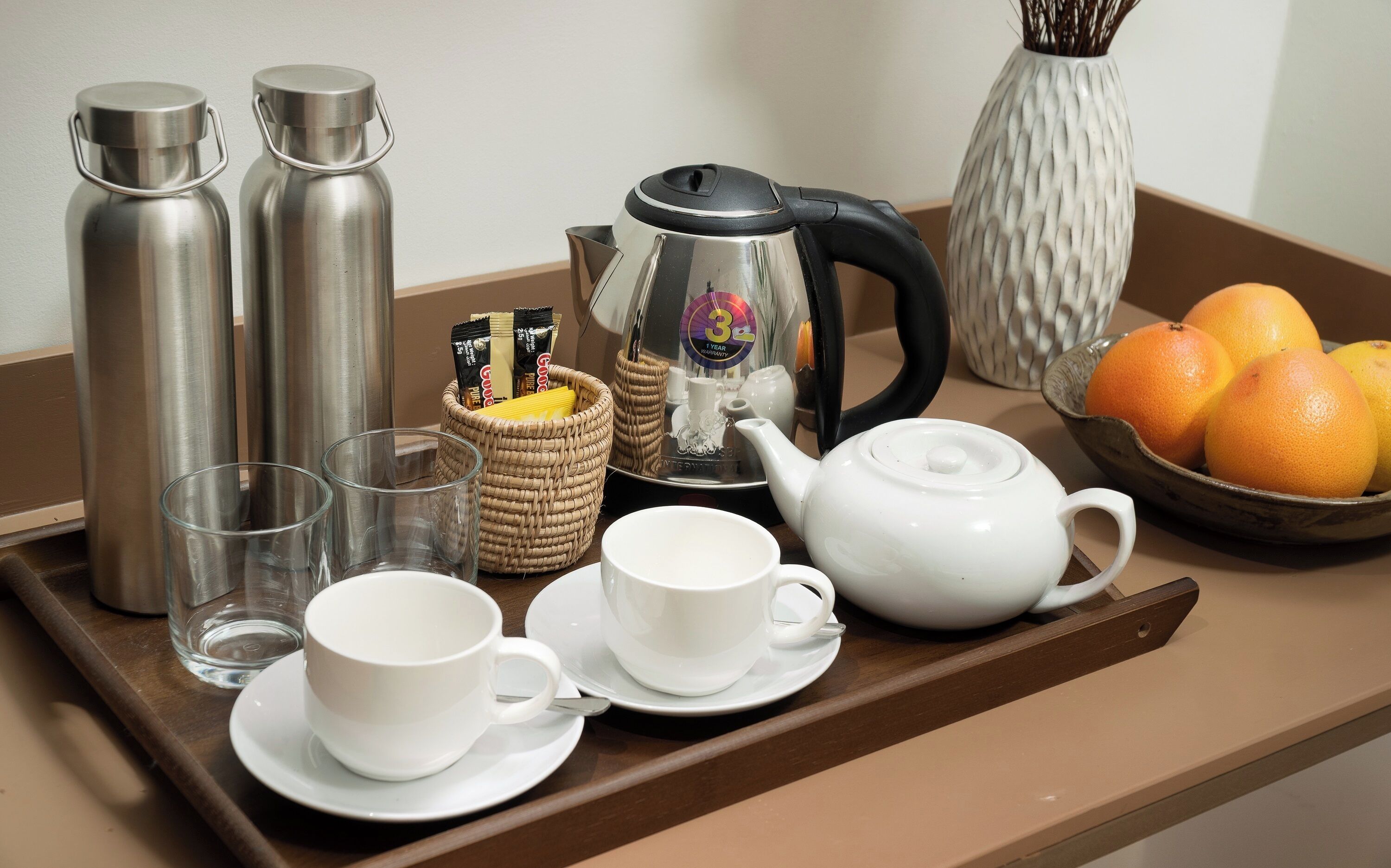 A tray with a teapot, two cups, two thermos flasks, a basket with tea bags, a vase with branches, and a bowl of oranges.