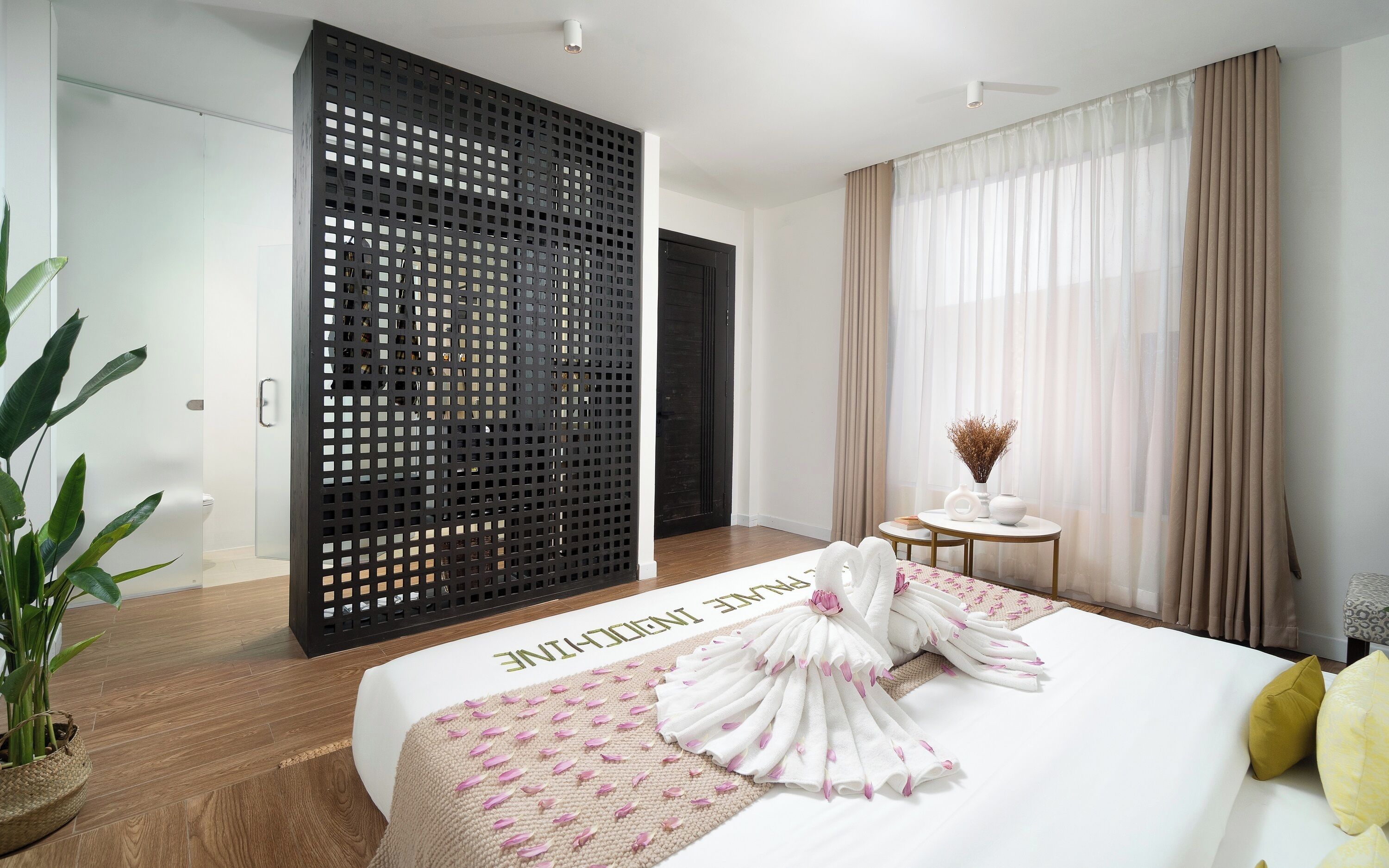 A hotel room with a bed, a small round table with a vase, a potted plant, and a sliding door.