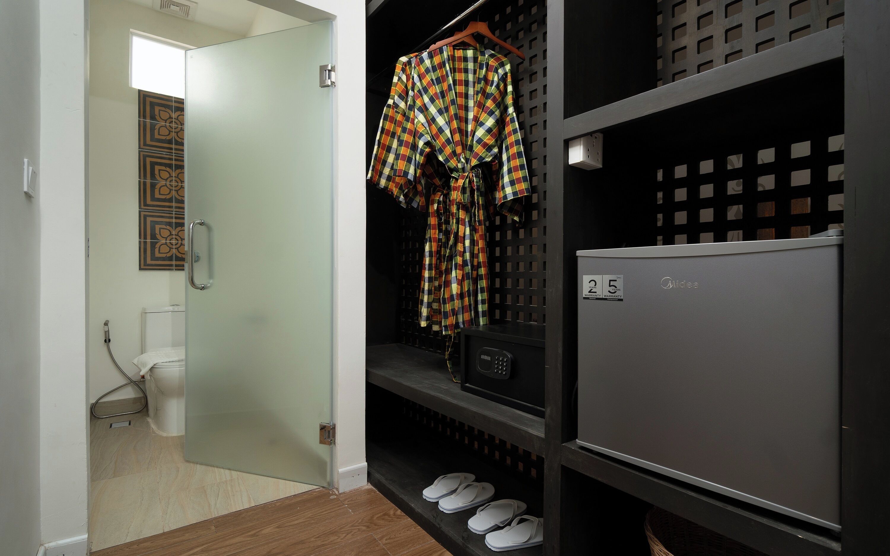 A hallway with a glass door, a white toilet, a black refrigerator, and a shelf with white slippers.