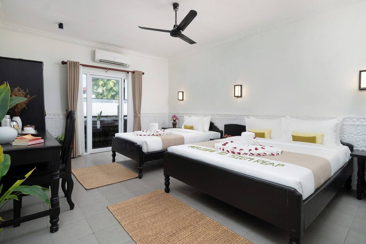 Habitación de hotel con dos camas, ventilador de techo, aire acondicionado, zona de comedor con mesa y sillas, y vista a través de una puerta de cristal.
